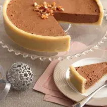 fotografia em tons de cinza e marrom de uma bancada cinza, contém um suporte para servir sobremesa transparente e redondo com uma torta com mousse de chocolate. Ao lado um prato rendo e bege com uma fatia da torta e um garfo ao lado para servir.