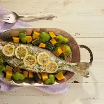 Fotografia em tons de lilás em uma bancada de madeira clara com um pano lilás, uma panela de ferro com o peixe assado com legumes dentro dela.