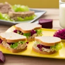 Fotografia de um apoio amarelo grande e retangular apoiado em uma mesa de madeira. Sobre o apoio tem três sanduíches de atum com beterraba e alface.