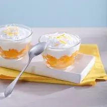 Fotografia em tons de amarelo em uma bancada de madeira clara, um pano amarelo, um recipiente branco retangular com duas taças de vidro com a sobremesa de laranja com creme de coco dentro delas.