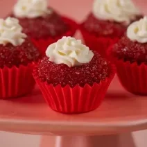 Fotografia de cinco unidades forminhas de cor vermelha, um junto da outra, e dentro de cada forminha tem um brigadeiro de red velvet, passado no açúcar cristal com uma decoração de cream cheese. Os brigadeiros estão sobre um apoio decorativo rosa alto.