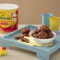 Fotografia em tons de amarelo e vermelho com bandeja azul ao centro. Em cima da bandeja existe um prato fundo com vários cookies feitos de NESTLÉ Aveia em Flocos e NINHO Primeira Infância