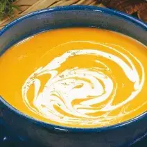 fotografia em tons de laranja, azul e marrom de uma bancada marrom vista de cima, contém um prato redondo azul com um recipiente redondo e azul com a sopa cremosa de cor laranja.