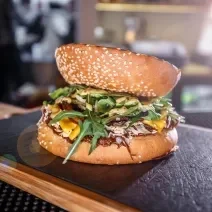 Fotografia de um hamburguer de carne e champignon temperado e servido com rúcula. A fatia do pão na parte de cima, está ao contrário, e o lanhe está sobre uma tábua de madeira com fundo preto.
