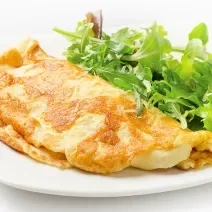 Fotografia de um omelete com crosta de queijo crocante dobrado ao meio, ao lado de uma salada de folhas, sobre um prato de vidro branco. O prato está sobre uma mesa branca.