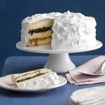 Fotografia em tons de branco e azul, ao fundo, sobre bancada azul, uma boleira de porcelana com bolo fatiado coberto com marshmallow e recheio de ameixa, prato de sobremesa com bolo de ameixa, dois pratos com talheres e um guardanapo com uma espátula.