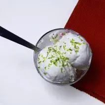 Fotografia em tons de vermelho e branco de uma bancada branca vista de cima. Contém uma taça com uma bola de sorvete com raspas de limão e uma colher para servir e ao lado um pano vermelho.