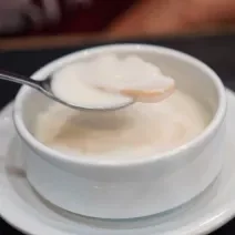 Fotografia de fatias de laranja cobertos por leite condensado dentro de um recipiente branco pequeno, sobre um prato branco pequeno.