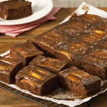 fotografia em tons de marrom e branco de uma bancada marrom vista de frente. Contém uma grade com um papel branco por cima, que contém um bolo de chocolate com pedaços de maçãs e cortado em quadrados