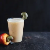 Fotografia de um copo de vidro com refresco de Neston com suco de caju, ao lado de um caju in natura sobre uma mesa escura e um fundo preto.