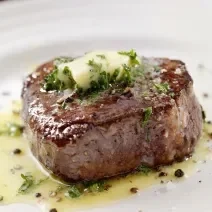 Fotografia de um bife mais alto feito, em um prato branco, com um azeite de ervas e pimenta sobre a carne e escorrendo no prato. Em cima da carne, manteiga e salsinha.