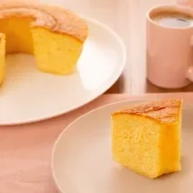 Fotografia de um prato raso branco grande com um bolo quase inteiro de fubá, na frente tem outro prato, menor e de tom mais escuro, com uma fatia do bolo de fubá. Um pouco mais ao fundo tem uma xícara rosa com café.