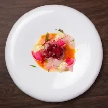 Fotografia em tons de branco com um prato branco ao centro. Em cima do prato existe uma receita de crudo de peixe de cor laranja e rosa, acompanhada de pedaços de limão