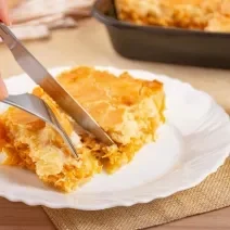 Fotografia de uma fatia de trota de frango em um prato raso de vidro branco. Tem uma mão masculina cortando um pedaço com garfo e faca. No fundo tem a uma forma preta com a torta assada.