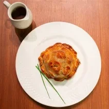 Fotografia de uma receita de pithivier paraense com duxelles de ora-pro-nóbis sobre um prato raso branco, o qual está sobre uma bancada de madeira. Ao lado do prato tem um recipiente fundo e pequeno com molho.