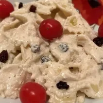 Fotografia de uma masssa farfalle cremosa com uvas passa e tomates cereja dentro de um prato branco de vidro.