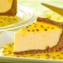 Fotografia de uma fatia grande de torta de maracujá com base de biscoitos e calda da fruta sobre e por volta da fatia, que está em um prato quadrado branco. No fundo da foto tem a torta inteira com mais calda de maracujá.