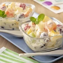 Fotografia de dois recipientes quadrados pequenos de vidro com salada de frutas e creme de maracujá com ninho em cada um, misturado. Por cima deles tem algumas folhas de hortelã, ao lado tem uma colher clara, também sobre um apoio retangular azul.