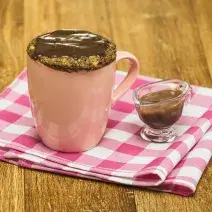 Fotografia em tons de rosa em uma bancada de madeira escura, um pano rosa xadrez, uma caneca rosa com um bolo de caneca de banana com mel, aveia e cobertura de chocolate. Ao lado, um potinho de vidro com a calda de chocolate.