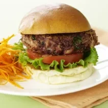 Fotografia de um hambúrguer com carne bovina, salada, ovo e queijo, acompanhado de cenoura cozida. O hambúrguer está sobre um prato branco que está apoiado em um pano cor de salmão. Tudo está sobre uma mesa verde claro.