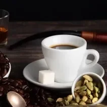 Fotografia em tons de marrom com uma xícara branca ao centro. Dentro da xícara existe um café acompanhado com um quadrado de açúcar. Ao lado existe uma vasilha com grãos de café.