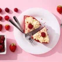 Fotografia vista de cima de um prato raso grande branco de vidro com duas fatias de torta red velvet, com dois garfos juntos ao lado. Ao redor do prato tem mais dois garfos e muitas frutas vermelhas.