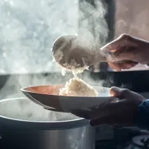 Fotografia de uma mão feminina colocando com uma colher de pau um arroz cremoso ao champanhe em um prato fundo. A imagem está contra a luz do sol, e se caracteriza pelo vapor saindo da panela.