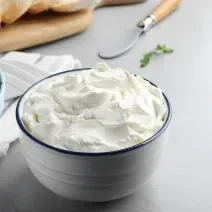 Fotografia em tons de branco com um pote branco ao centro. Dentro do pote existe uma receita de cream cheese caseiro, um creme branco feito com vinagre, leite e creme de leite.