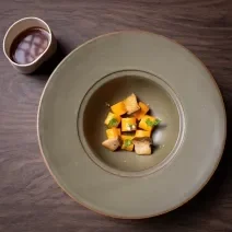 Fotografia vista de cima de um prato escuro com o meio fundo. Dentro do prato tem uma receita de nhoque de batata doce laranja, dashi de missô e cogumelos tostados. O prato está sobre uma bancada de madeira escura.