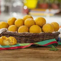 Fotografia em tons de marrom e vermelho em uma bancada de madeira com uma cesta oval de vime e vários bolinhos de abóbora assados com queijo. Ao lado, tomates inteiros e um pedaço de tomate.