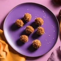 Fotografia em tons de roxo com um prato roxo ao centro. Dentro do prato existe pequenas trufas de cenoura com uva passa cobertas com coco ralado