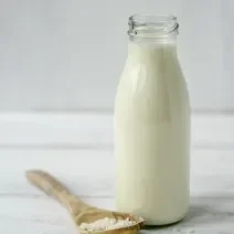 Fotografia de uma bebida de limão com leite MOÇA e neston em uma garrafa de vidro aberta sobre uma mesa de madeira branca. Ao lado tem uma colher de madeira com sementes brancas.