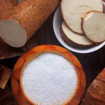 fotografia em tons de marrom e branco de uma bancada marrom vista de cima, contém um recipiente redondo marrom com a farinha dentro e ao lado um recipiente redondo branco com pedaços de mandioca. Em volta contém mandiocas.