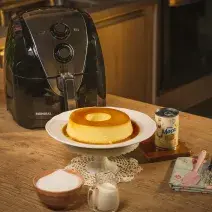 Fotografia em tons de amarelo em uma bancada de madeira, um suporte para bolo branco e o pudim e Leite Moça em cima dele. Ao fundo, uma air fryer Mondial, uma lata de leite Moça, um paninho azul florido com uma espátula rosa e um fouet em cima.