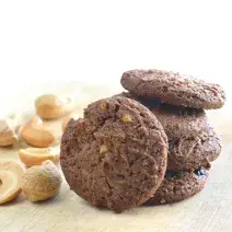 fotografia em tons de branco, marrom e bege de uma bancada bege vista de frente, ao centro biscoitos de chocolate e ao lado castanhas de caju