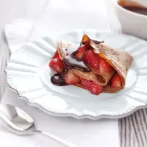 fotografia em tons de branco, marrom e vermelho de uma bancada cinza vista de frente, contém um prato branco redondo com um Crepe de Chocolate com Morangos e ao lado uma colher para servir.