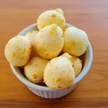 Fotografia de um ramekin branco com vários pães de queijo. O recipiente está sobre uma mesa de madeira.