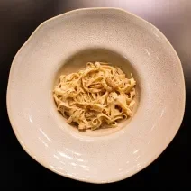 Fotografia em tons de cinza e preto com um prato cinza ao centro. Em cima do prato existe uma receita de cacio e pepe feita com macarrão tagliatelle e finalizado com pimenta preta moída