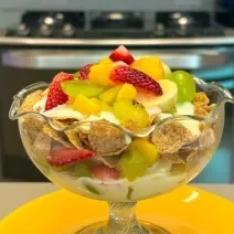 Fotografia em tons de amarelo com uma taça de vidro ao centro. Dentro da taça existe uma receita de salada de frutas com creme de Leite NINHO Adulto e Nesfit Cereal