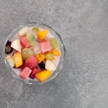 Fotografia vista de cima de uma taça de vidro com frutas sortidas servidas com Leite MOÇA sobre uma bancada de cimento queimado.