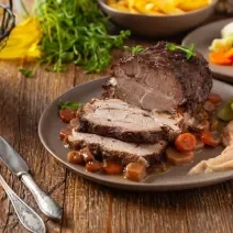 Fotografia de uma peça de cupim assada com manteiga sobre alguns vegetais sobre um prato raso cinza. Ao lado da carne tem talheres sobre uma mesa de madeira.