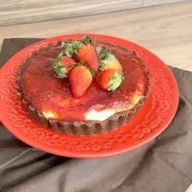 Fotografia em tons de vermelho com um prato vermelho ao centro. Em cima do prato existe um cheesecake de Galak com queijo gorgonzola, cobertura de morango e morangos inteiros