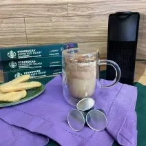 Fotografia em tons de verde e roxo em uma bancada de madeira, um paninho verde e roxo ao centro com uma xícara de vidro e o café tiramisu dentro. Ao fundo, uma máquina nespresso.
