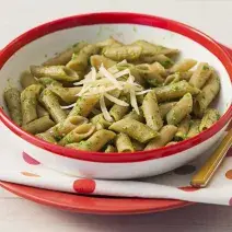 Fotografia em tons de verde, vermelho e branco, com prato branco com borda vermelha contendo macarrão e queijo ralado, garfo ao lado, sobre guardanapo branco com bolinhas coloridas e prato vermelho, tudo sobre bancada branca.