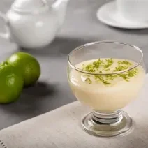 Fotografia em tons de branco com uma taça de sobremesa de vidro ao centro dentro da taça existe uma receita de mousse de limão coberta com raspas de limão.