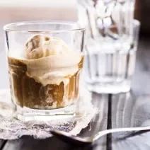 Fotografia de dois copos de vidro sobre uma mesa de madeira. O copo da frente tem uma bola de sorvete de creme com café e chocolate.