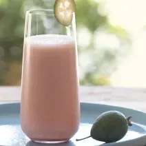 Fotografia de uma vitamina de goiaba com leite e neston com uma fatia fina de uma rodela de fruta na borda do copo de vidro. A bebida está apoiada em um prato azul, sobre uma mesa de madeira.