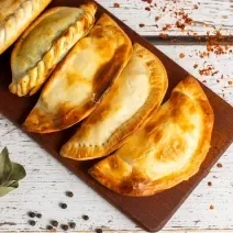 Fotografia vista de cima de cinco pasteis assados na air fryer sobre uma tábua retangular de madeira pequena, a qual está sobre uma mesa de madeira branca.