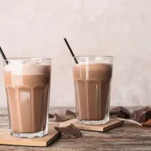 Fotografia de dois copos de vidro grandes com vitamina de nescau. Dentro dos copos, em cada um, tem um canudo preto e alguns cubos de gelo.