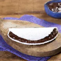 Fotografia em tons de marrom e roxo de uma bancada de madeira com um paninho roxo, sobre ele um tábua de madeira com uma tapioca. Ao fundo um recipiente roxo com creme de dark chocolate.